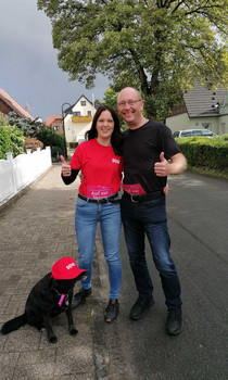 Ein Paar steht mit Hund auf einer regennassen Straße. Der Himmel ist bewölkt und wirkt dunkel.