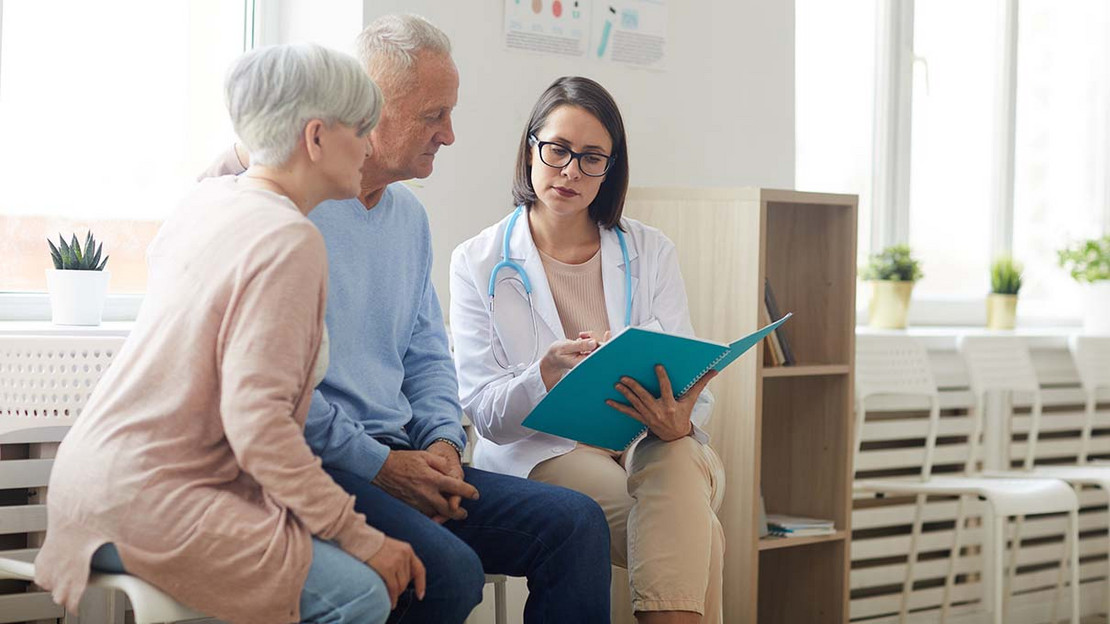 Eine Ärztin sitzt mit zwei älteren Personen auf Stühlen und zeigt ihnen etwas in ihren Unterlagen.