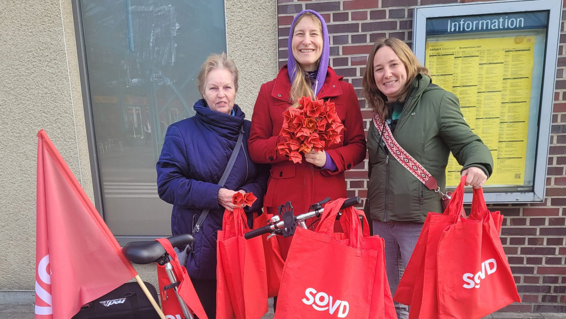 Drei SoVD-Frauen mit Papierrosen und Taschen vor dem Bremerhavener Hauptbahnhof