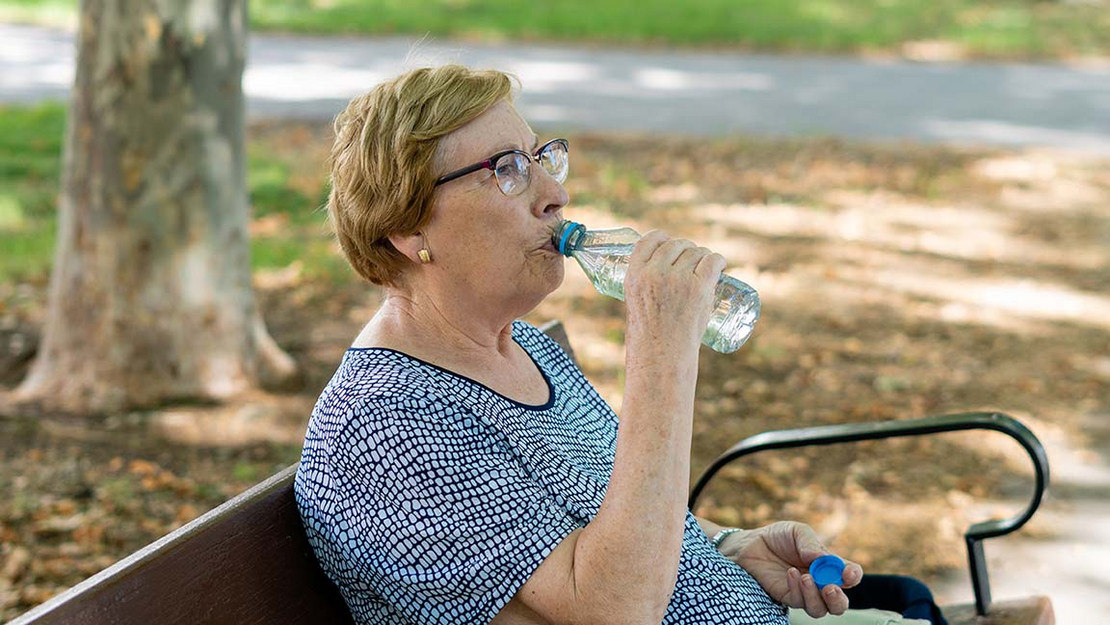 Eine ältere Frau sitzt auf einer Bank im Schatten eines Baumes und trinkt aus einer Wasserflasche.