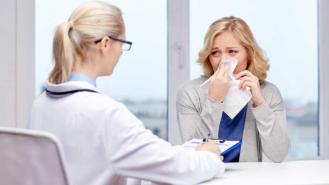 Ärztin mit einer Patientin, die ein Taschentuch vor die Nase hält. 