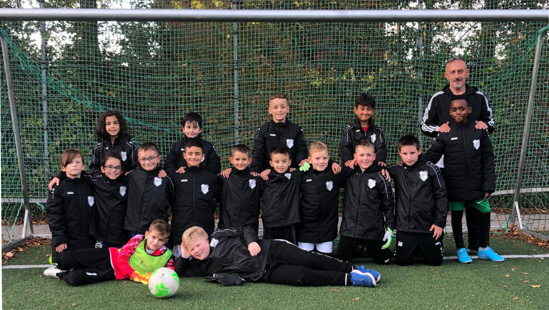 Die Fußballgruppe mit 16 jungen Kickern und ihrem Trainer mit schwarzem Fuß-ball-Outfit stehen auf dem grünen Rasen vor dem Tor